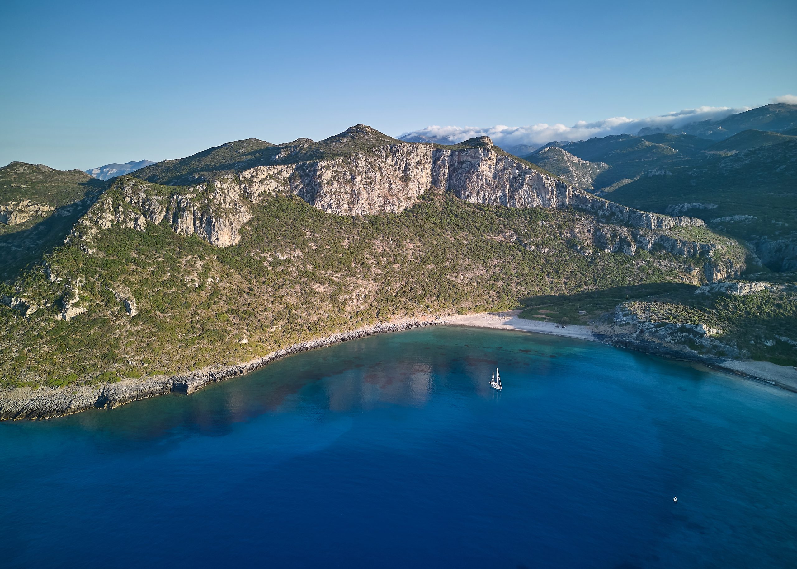 Aerial view of a sailing yacht anchored alone in a deep blue bay beneath limestone cliffs in Greece
